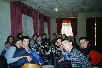 Group photo of young adults in a dimly lit room with red curtains, likely a 1990s social gathering. Wooden table filled with ...