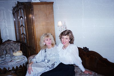 Two women pose indoors in a mid-century interior, likely 1970s–1980s. The woman on left wears a patterned blouse with volumin...