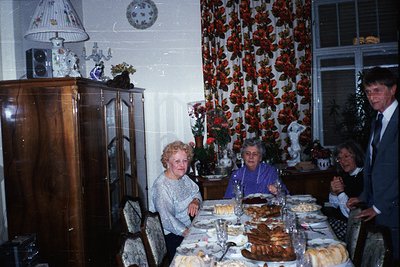 Vintage indoor gathering featuring four adults seated at a long dining table adorned with tiered cakes, floral arrangements, ...