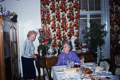 Vintage indoor scene featuring two elderly women in a mid-century dining setting. One woman stands by a floral-patterned curt...