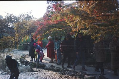 A group of people in autumn foliage, likely a Buddhist temple or monastery setting. Monks in maroon robes lead children and a...