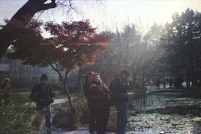 Vintage 35mm shot of a park scene with low-angle sunlight filtering through autumn foliage. Four people—two adults and two ch...