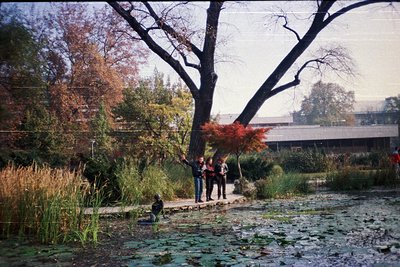 Vintage 1970s-style park scene with three people standing near a pond. Autumn foliage dominates, with a prominent tree displa...