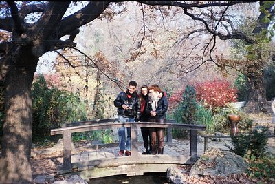 Three individuals pose on a modern glass-railing bridge in a park during autumn, surrounded by foliage in warm hues. The man ...