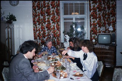 Vintage indoor family gathering at a festive holiday table, 1970s-1980s. Decorated with floral wallpaper, a lit Christmas tre...