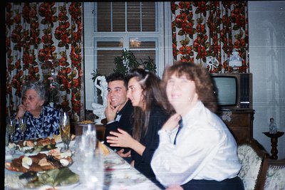 Vintage indoor gathering featuring four adults seated at a table laden with food and drinks. Decor includes red floral wallpa...