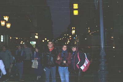 Urban night scene with three individuals posing near a crosswalk. Reflective glass surface obscures background details. Peopl...