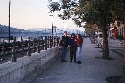Three individuals pose on Budapest’s Danube Promenade, flanked by ornate black wrought-iron railings and mature trees. The ic...