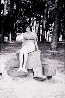 Vintage black-and-white portrait of a woman in a sleeveless swimsuit pose on tree stump seating, surrounded by dense forest. ...