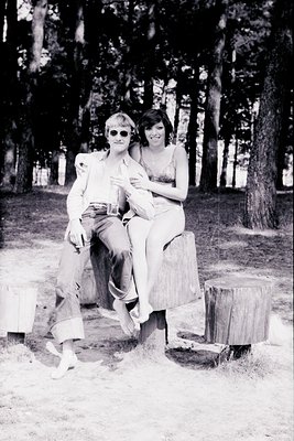 Two individuals pose casually on tree stumps in a wooded area, 1970s style. Man wears sunglasses, a light jacket, and rolled ...