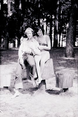 Couple posing affectionately on tree stumps in a wooded area, mid-20th century. Man in rolled-sleeve shirt and jeans, woman i...