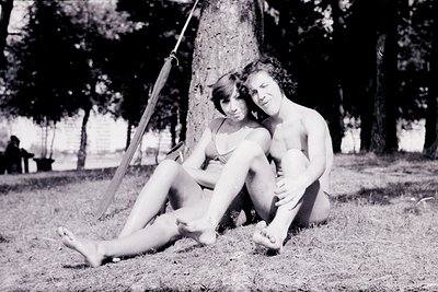 Black-and-white photo of two individuals sitting on a grassy park swing, leaning against a tree trunk. Mid-20th century attir...