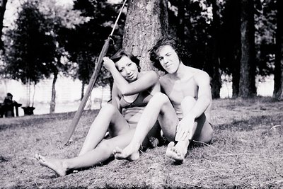 Mid-20th century black-and-white photo of two individuals swinging on a rope tied to a tree in a park. Woman in a sleeveless ...