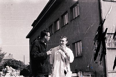 Two men stand near a building’s exterior wall, appearing to examine structural damage. One wears glasses and a dark jacket; t...