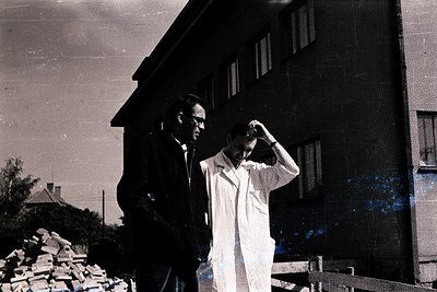 Two men stand outdoors, one in a dark suit and glasses, the other in a white lab coat, appearing to discuss something. A larg...