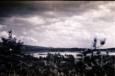 A panoramic landscape features a wide body of water reflecting a dramatic, cloudy sky. Dense foliage frames the lower portion...
