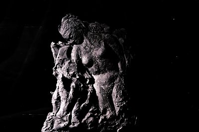 A textured, monochromatic sculpture depicts a figure in a contemplative pose, likely female. The work features layered, rough...