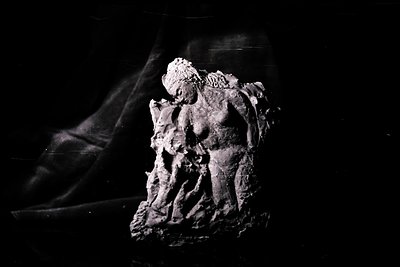 A textured, grayscale sculpture of a female figure, seemingly emerging from a rocky base. The form appears abstract, with det...