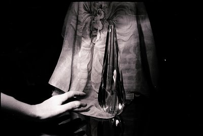 A clear, elongated glass vase sits on a reflective surface, partially obscured by a draped, textured fabric. A hand gently to...