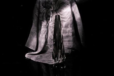 Elegant, angular black glass vase rests on draped fabric. High contrast black and white photograph with visible grain. Likely...