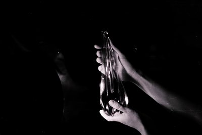 A black and white photograph shows two hands delicately holding a clear glass bottle, illuminated by a dramatic light source ...