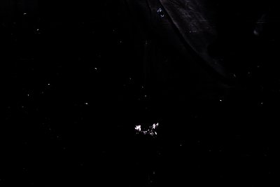 A minimalist, high-contrast image depicting a small, geometric sculpture or mobile suspended in darkness. The object features...
