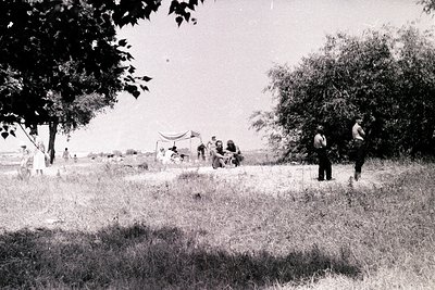 A vintage black-and-white photograph depicts a group gathered outdoors, likely a leisure or picnic scene. A makeshift shelter...
