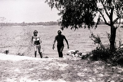 Monochrome photograph depicts a lakeside scene. A woman in a bikini stands on a sandy shore, facing a man wading into the wat...