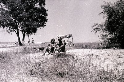 A black and white image depicts two men seated in tall grass, appearing relaxed, with a wider group visible in the distance b...