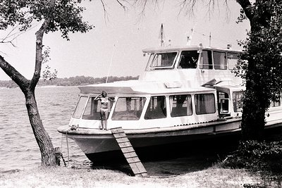 A two-story passenger ferry docks at a grassy shoreline. A young person stands on the vessel’s walkway near a wooden staircas...