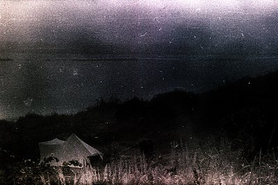 Night scene captures a simple, peaked-roof structure nestled among tall grasses, set against a dark hillside under a starry s...