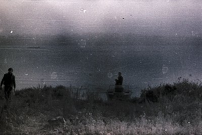 A striking, grainy black and white photo captures two figures against a hazy, expansive coastline. One person walks away from...