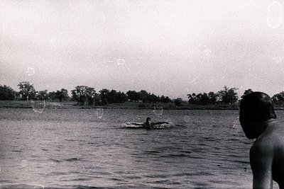 A swimmer appears mid-stroke in a still body of water. A figure, likely the photographer, stands in the foreground, viewed fr...