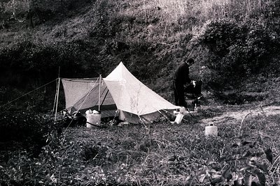 A lone figure stands near a small, weathered tent pitched on a grassy slope. Camping equipment, including a barrel and stacke...