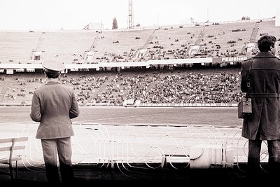 Two men stand overlooking a large stadium filled with spectators. One, in a military-style uniform & cap, faces forward. The ...