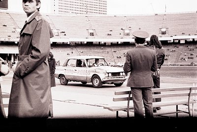 Monochrome image depicting an "Auto-Rodeo" promotional event within a large, tiered stadium. Two uniformed men stand observin...