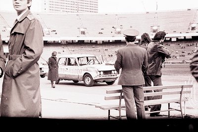 A 1970s black and white image shows a man in a trench coat, alongside uniformed officials observing a woman near a Lada autom...