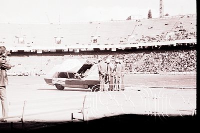A black and white photo shows a vintage sedan, likely 1960s-70s, with its hood raised, surrounded by uniformed men on a stadi...