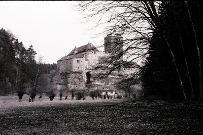 Striking, austere view of a medieval castle dramatically perched atop a rocky crag. Extensive fortifications & a tower define...