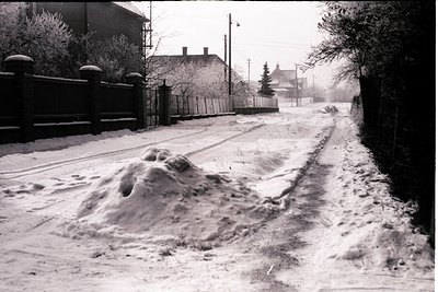 Snow-covered road bisects a residential scene. A large snowdrift dominates the foreground, while a wooden fence and distant h...