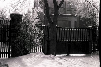 A gate stands partially open in a snow-covered yard, facing a large, multi-windowed house. A stone pillar and a dense, froste...