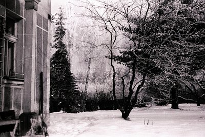 A stark black and white image depicting a snow-covered courtyard, likely in Eastern Europe. A multi-story building with visib...