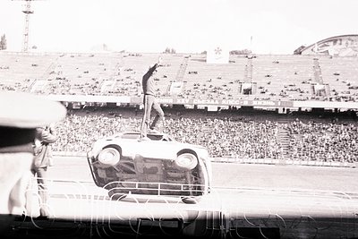 Man stands atop an overturned car in a stadium, raising his hand. Large crowd visible in bleachers. Cyrillic text on stadium ...