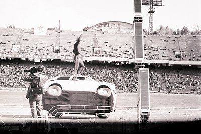 A lone figure stands atop an overturned car within a large stadium, appearing to gesture or wave to a crowd. A photographer c...