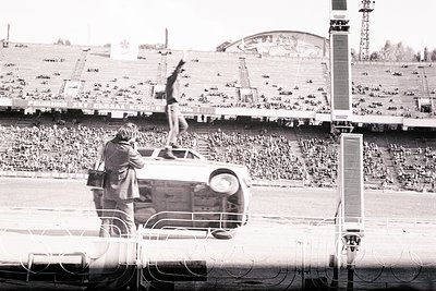 Man stands atop an overturned car during a public spectacle amidst a large stadium crowd. A photographer documents the scene....