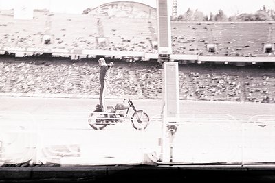 A man balances precariously atop a vintage motorcycle in a large stadium. The setting appears to be a sports arena, with tier...