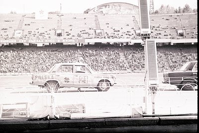 A 1960s-70s rally car, labeled "POLSKA," speeds across a track during an event. A large crowd fills the stadium bleachers. A ...