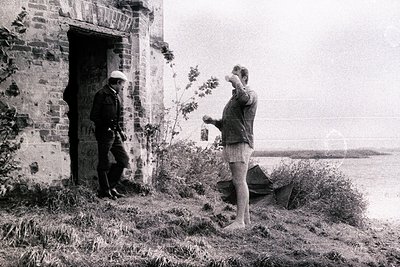 Two men near a weathered stone structure overlooking a body of water, likely a lake or sea. One man, in a cap and jacket, lea...