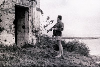 A barefoot man in a robe and shorts stands beside a crumbling brick structure overlooking a body of water. The building appea...