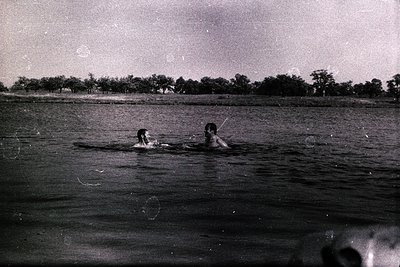 Two individuals float in a tranquil body of water, partially submerged, with a dense treeline forming the distant backdrop. A...